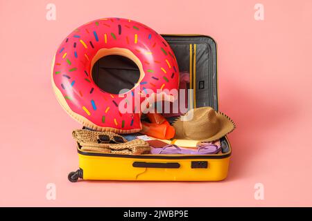 Offener Koffer mit Kleidung, verschiedenen Strand-Accessoires und Reisepass auf rosa Hintergrund Stockfoto