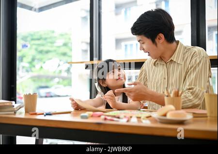 Lächelnder und fröhlicher asiatischer Kunstlehrer lehrt und unterhielt sich mit seinem jungen kleinen Mädchen im Café. Stockfoto