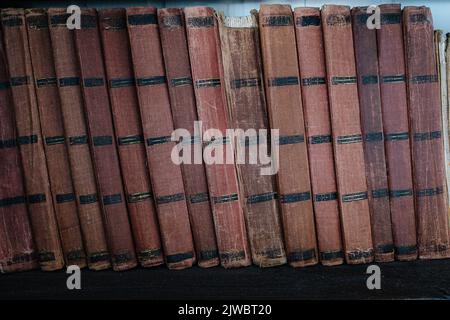 Alte Bücher in einer Reihe mit namenlosen Stacheln und zerlumpten, abgenutzten Bezügen auf einem Regal. Dunkelbraun mit schwarzen Streifen. Stockfoto