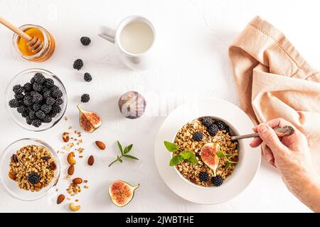 Die Hand einer jungen Frau mit einem Löffel Müsli oder Müsli gefüllt während des Frühstücks. Ein Teller mit Müsli, Brombeeren und eine Scheibe Feigen. Draufsicht Stockfoto