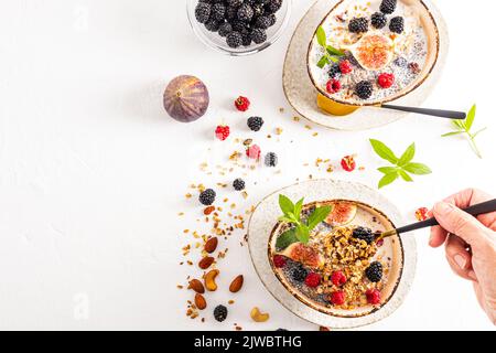 Frühstück für zwei Personen. Gekochtes köstliches Müsli in Keramikschalen mit Nüssen, Beeren, Minze und Feigen. Eine Frauenhand hält einen Löffel Müsli Stockfoto