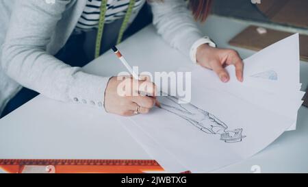 Die Nahaufnahme des Modedesigners zeichnet Linien auf der Skizze der Kleidung. Die Frau ist beschäftigt und konzentriert sich auf die Arbeit. Stockfoto