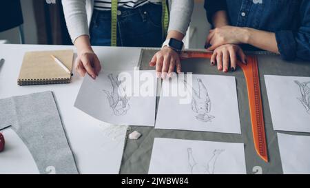 Nahaufnahme der gepflegten Hände der Schneider, die auf dem Nähtisch durch Schwarz-Weiß-Skizzen gehen. Junge Frauen diskutieren über Bekleidungsdesigns. Kreatives Schneiderkonzept. Stockfoto