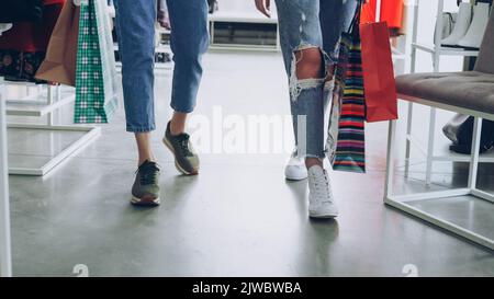 Die Beine der Frauen gehen langsam durch den luxuriösen Laden. Frauen tragen Jeans und Sportschuhe und tragen helle Papiertaschen. Shopaholic Lifestyle-Konzept. Stockfoto