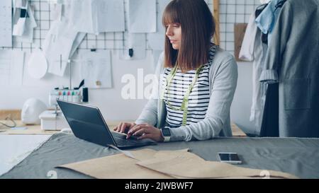 Weibliche Bekleidungsdesign-Unternehmerin nutzt Laptop, um mit ihren Kunden zu kommunizieren und hergestellte Kleidungsstücke online zu verkaufen. Modernes E-Commerce im Small Business-Konzept. Stockfoto