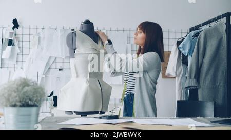 Junge attraktive Näherin misst sorgfältig Schneiderpuppe mit Klebeband, um neue Kleidung mit diesen Maßen zu machen. Arbeitsreichen Tag in Kleidung Workshop-Konzept. Stockfoto