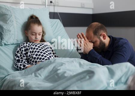 Krankes kleines Mädchen unter medikamentöse Behandlung ruht in der Kinderklinik, während die Eltern für ihre Gesundheit beten. Besorgter Vater, der für eine kranke Tochter betet, die auf dem Patientenbett liegt Stockfoto