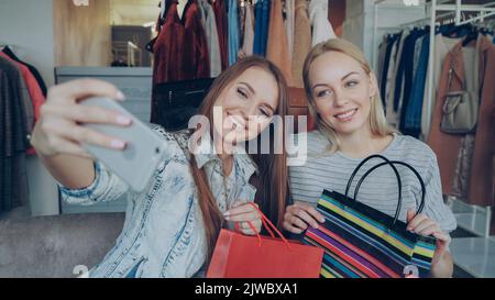 Nahaufnahme von zwei jungen Frauen, die Selfie mit dem Smartphone machen, nachdem sie im Bekleidungsgeschäft für Frauen einkaufen waren. Zuerst posieren sie mit Papiertüten und lachen, dann schauen sie sich Fotos auf dem Bildschirm an Stockfoto