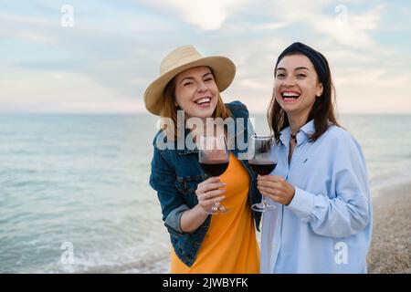 Zwei junge hübsche Frauen mit Rotwein am Abend am Meer bei Sonnenuntergang. Stockfoto