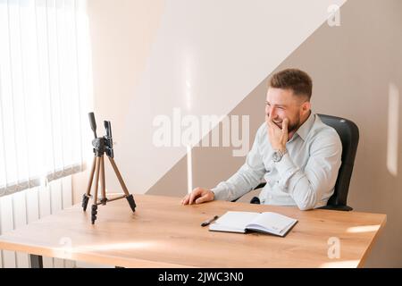 Ein junger bärtiger Geschäftsmann, der im Büro arbeitet und am Tisch sitzt, der ein Smartphone für ein Online-Meeting verwendet, um sich Notizen in einem Tagebuch zu machen. Freiberufler arbeitet Remote. Der Student bereitet sich auf das Seminar vor. Online-Marketing Stockfoto