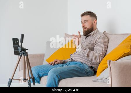 Lächelnder bärtiger Kerl, der online mit einem Smartphone auf einem Stativ ein Video dreht, während er auf dem Sofa im Wohnzimmer sitzt und Hemd und Jeans trägt. Einfluss sozialer Medien, Erstellung von Inhalten und Online-Beratung Stockfoto