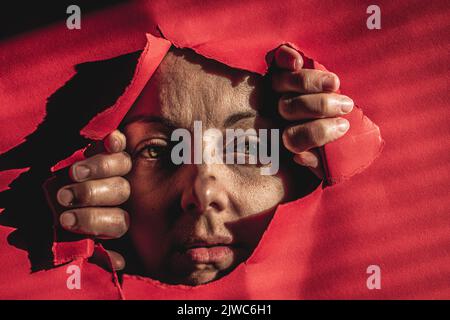 Porträt einer traurigen Frau, die durch ein Loch in Papier auf die Kamera schaut. Ich möchte freie Idee sein. Nahaufnahme Porträt einer verängstigten kaukasischen jungen Frau. Stockfoto