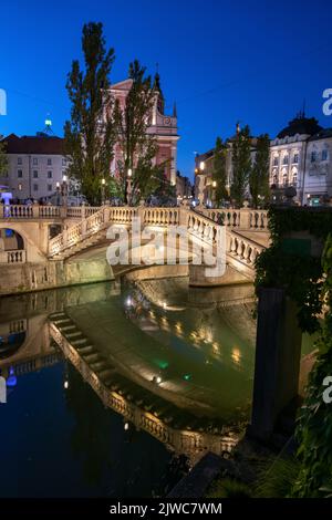 Stadt Ljubljana in Slowenien, Dreifachbrücke (Tromostovje) mit Spiegelung im Fluss Ljubljanica bei Nacht und Franziskanerkirche der Verkündigung. Stockfoto