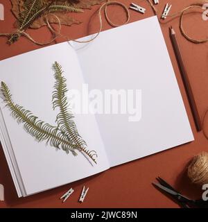 Getrocknetes gepresstes Blatt auf Papier aus Notizbuch mit Bleistift, Schere und Garn auf braunem Hintergrund. Hobby, Handarbeit, Blumenkunst und Boho-Konzept. Quadratisch Stockfoto