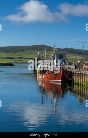 Dingle, Irland - 7. August 2022: Rotes Fischerboot auf den Docks von Dingle Harbour in der Grafschaft Kerry mit Reflexionen im ruhigen Wasser Stockfoto