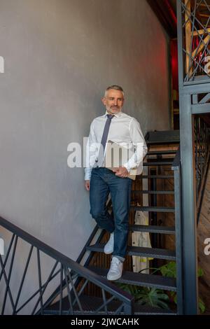 Eleganter, attraktiver grauhaariger Mann mit dem Computer, der nach unten geht Stockfoto