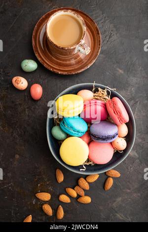 Bunte Makronen und Schokoladeneier in Keramikschale, Tasse Kaffee auf schwarzem Betongrund. Draufsicht, flach liegend, Nahaufnahme, Stillleben. Pause Stockfoto