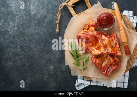 Schweineribs. Rohes Fleisch. Rohe Schweinefleischrippen in Marinade mit Gewürzen, Rosmarin, Tomatensauce und Knoblauch auf altem Holzständer auf schwarzem Steintisch Hintergrund. Bar Stockfoto