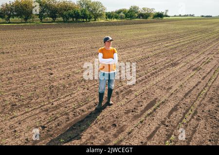 Luftaufnahme einer Farmerin, die auf dem Maiskeimfeld steht und Feldfrüchte untersucht. Landarbeiter mit Truckerhut und Jeans auf Plantage von Drohne p Stockfoto