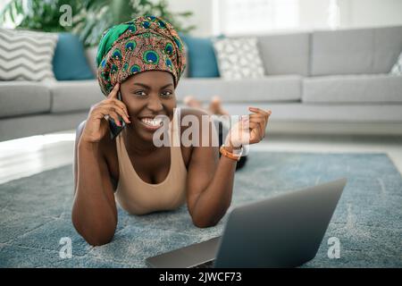 Schöne afrikanische Frau zu Hause, lächelnd und lachend in die Kamera schauend, mit Handy, das traditionelle Kopftuch tragend, überrascht und happ Stockfoto