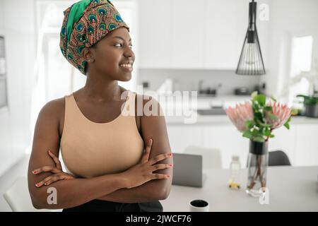 Schöne afrikanische Frau zu Hause, lächelnd, tragend traditionelles Kopftuch und Blick in die Ferne mit Kopieplatz Stockfoto