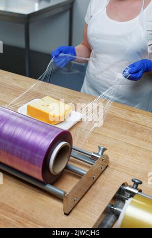 Lebensmitteleinzelhändler. Lebensmittelverpackungen. Nicht erkennbare Arbeiterin in blauen Einweghandschuhen wickelt Stück Käse mit transparenter Folie, die sie zieht f Stockfoto