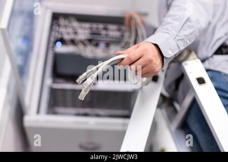 Server-Hardware-Setup. Ein männlicher Mitarbeiter konfiguriert das lokale Netzwerk, indem er Kabel und Stecker festhält, um eine Verbindung zum Server herzustellen. Nahaufnahme des Steckers in der Hand eines stehenden Mannes Stockfoto