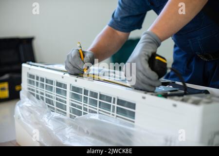 Männlicher Techniker macht beim Messen der Klimaanlageneinheit mit Bleistift Spuren. Nahaufnahme von Menschenhand in Schutzhandschuhen hält Bleistift und markiert mich Stockfoto