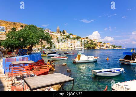 Symi, Griechenland - 26. August 2022: Panoramablick, Skyline der kleinen Oase der Insel Symi. Dorf mit winzigen Strand und bunten Häusern auf Felsen. Gipfel der Berge an der Küste von Rhodos Stockfoto