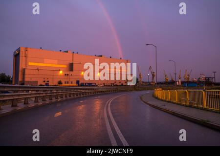 Regenbogen im Himmel bei Sonnenuntergang über einer Straße, die durch ein Industriegebiet führt Stockfoto