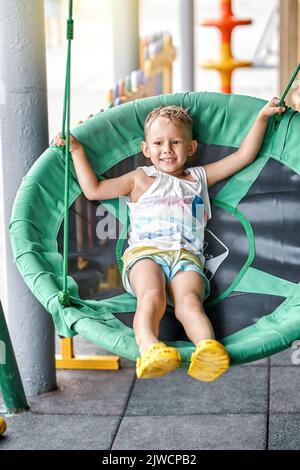 Der kleine blonde Junge sitzt in einem grünen Schaukelstuhl im Küstenresort. Kleine Kinder schaukeln auf dem Kinderspielplatz des Hotels und genießen einen schönen Urlaub am Meer Stockfoto