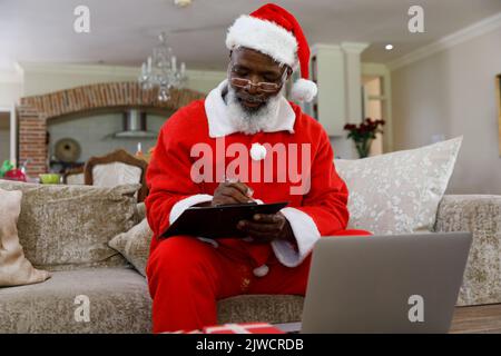 Älterer afroamerikanischer Mann, der ein Kostüm des weihnachtsmannes trägt und auf der Zwischenablage schreibt Stockfoto