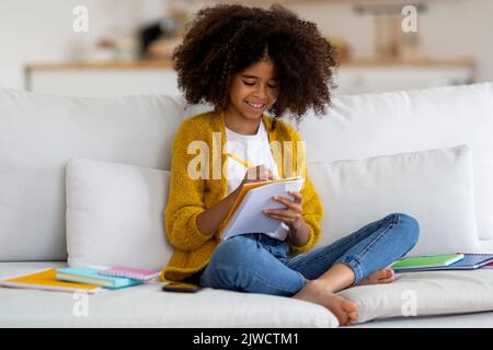 Glückliches afroamerikanisches Schulmädchen, das auf der Couch sitzt und Essay schreibt Stockfoto