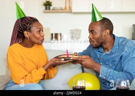 Glückliches schwarzes Paar in Partyhüten, das Kerzen auf Geburtstagskuchen bläst, Wein trinkt und gemeinsam den Geburtstag feiert Stockfoto