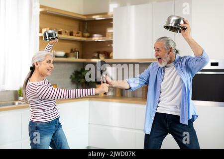 Fröhliche ältere Ehepartner, die gemeinsam in der Küche Spaß haben und spielerisch mit Küchengeräten kämpfen Stockfoto
