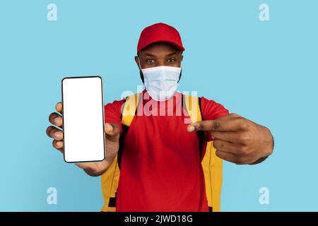 Schwarzer Junger Delivery Guy In Medizinischer Maske Zeigt Auf Unbeschriftem Smartphone Stockfoto