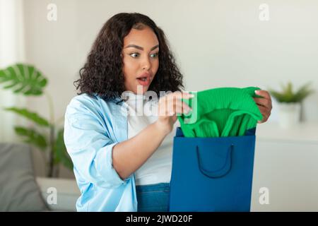 Erstaunt Afroamerikanische Frau Auspacken Einkaufstasche Mit Kleidung Im Innen Stockfoto