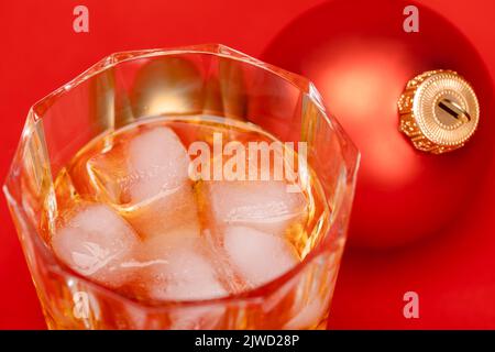 Glas Whiskey mit Eiswürfeln und weihnachtskugel auf rotem Hintergrund - weihnachtsgetränk-Konzept Stockfoto