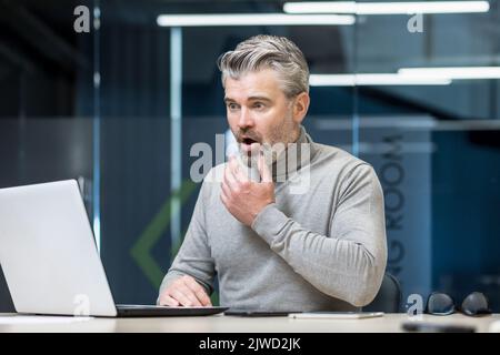 Älterer grauhaariger Geschäftsmann war schockiert, als er schlechte Nachrichten las, überrascht und verärgert reifer erfahrener Mann, der mit einem Laptop im modernen Bürogebäude arbeitete. Stockfoto