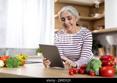 Porträt Einer Glücklichen Älteren Frau, Die Beim Kochen In Der Küche Ein Digitales Tablet Verwendet Stockfoto