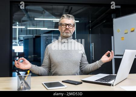 Ein grauhaariger Geschäftsmann, der im Bürogebäude meditiert, sitzt auf einem Stuhl am Schreibtisch, ein reifer Mann trägt eine Brille mit geschlossenen Augen in Lotusposition, ein Chef arbeitet am Arbeitsplatz mit einem Laptop. Stockfoto