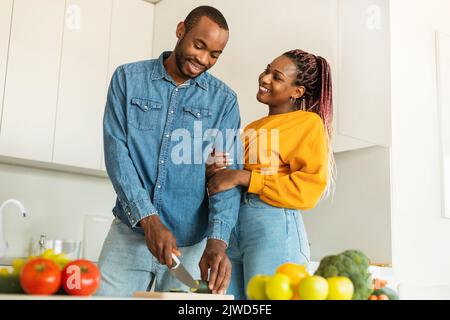 Liebevoller junger schwarzer Mann, der köstliches Abendessen für seine schöne Frau kocht, Gemüse schneidet, helles Kücheninnere Stockfoto