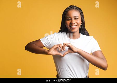 Lächelnd attraktive Brünette Frau schaut auf die Kamera und zeigt Herzform mit ihren Handflächen, gestikulende Liebe, romantisches Stimmungskonzept, isoliert auf gelbem Hintergrund Stockfoto