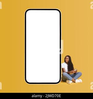 Multirassische Frauen surfen im Internet auf dem Smartphone, chatten in sozialen Netzwerken, verwenden mobile Apps, schreiben online mit Freunden, teilen Nachrichten, während sie in der Nähe eines riesigen Smartphones mit leerem leeren Bildschirm sitzen Stockfoto