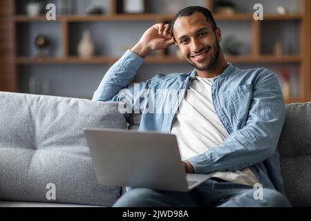 Schöner Afroamerikanischer Mann Mit Laptop, Während Er Auf Der Couch Zu Hause Sitzt Stockfoto