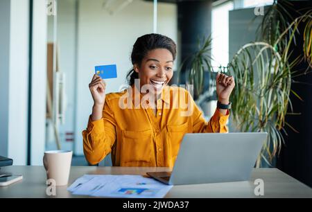 Überglücklich, schwarze Büroangestellte, die die Kreditkarte vor dem Laptop hält und eine „JA“-Geste macht und am Arbeitsplatz sitzt Stockfoto
