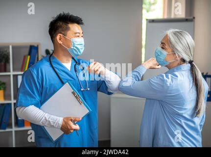 Lächelnder erwachsener chinesischer Mann, Arzt in Schutzmaske, begrüßt die alte Patientin in der Klinik mit Ellbogen Stockfoto