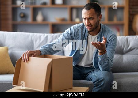 Falsches Paket. Verwirrter schwarzer Mann, der zu Hause einen offenen Karton ansieht Stockfoto