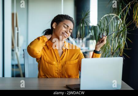 Nackenschmerzen. Schwarze Geschäftsfrau massiert schmerzenden Hals, leidet unter Schmerzen nach der Arbeit am Computer, sitzt im Büro Stockfoto