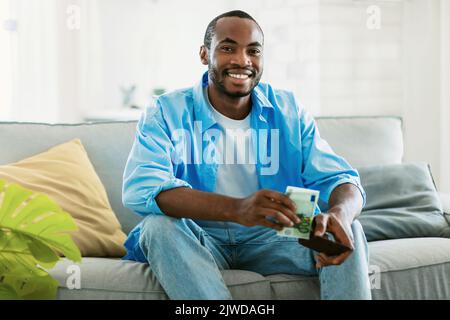 Positiver afroamerikanischer Mann, der Geldbörse hält und Geld herausnimmt, zu Hause auf dem Sofa sitzt und die Kamera anlächelt Stockfoto